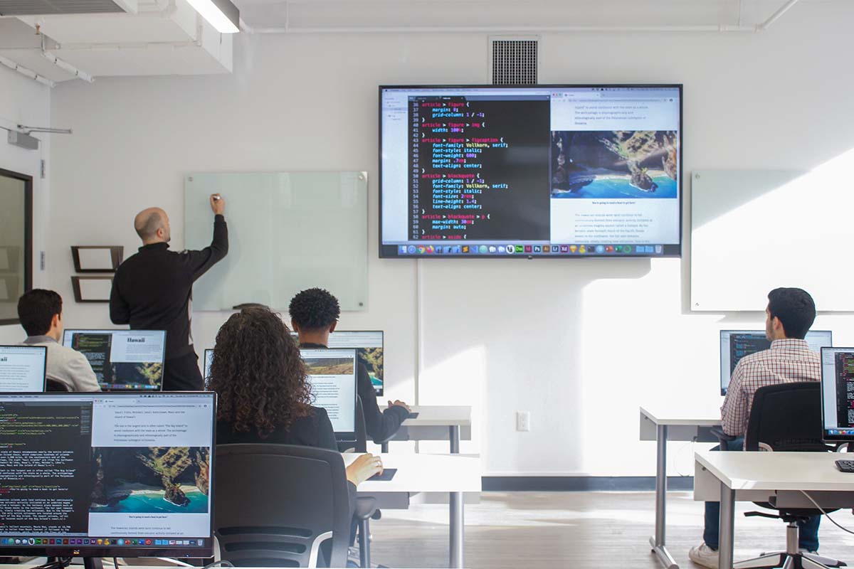 Students seated in computer classroom with workbooks, paying attention to instructor